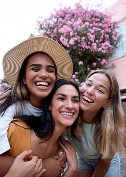 Vertical. Three cheerful female friends taking selfie laughing while enjoying their vacation on a picturesque street adorned with vibrant pink flowers, capturing moments of joy and togetherness