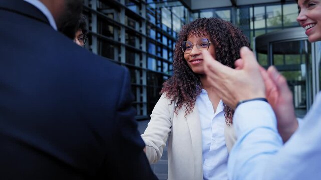 Black Latina professional shaking hands with a male colleague outside a modern office while coworkers gather around. Corporate scene conveying networking, diversity and professional collaboration