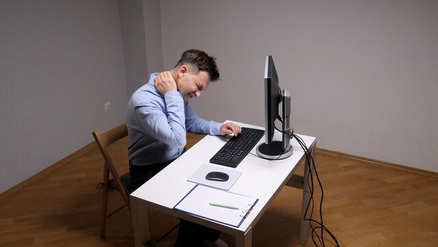 Man experiencing acute neck and shoulder pain while working at a computer in a home office, highlighting issues of poor posture, stress, and discomfort from prolonged desk work
