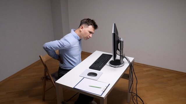 Man sitting at a desk and rubbing his lower back, feeling discomfort and strain while working on a computer, highlighting issues related to sedentary office work and poor posture