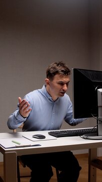 Stressed man sitting at a white desk in a dimly lit office, looking at a desktop computer monitor with an expression of anger and frustration, gesturing with his hands