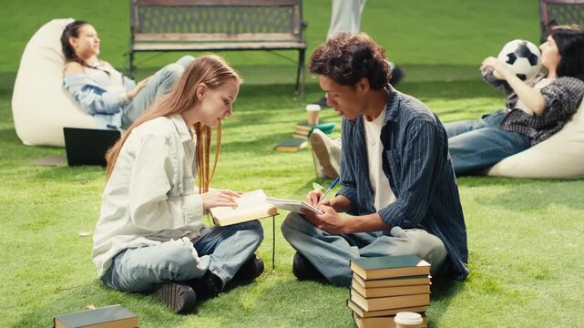University students celebrate teamwork with a high five on an outdoor campus lawn, smiling beside a laptop with notebooks and books after successful collaboration during a group study session.