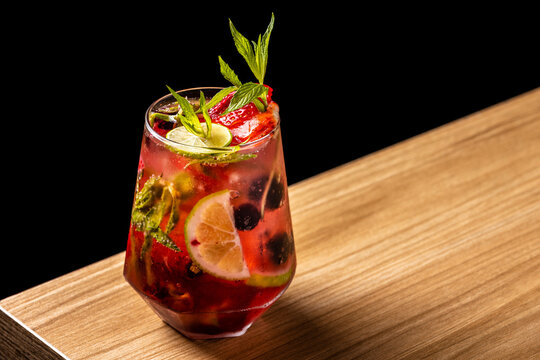Fruity berry smash cocktail with blackberries, strawberries, mint, and lime on a wooden bar surface with dark background
