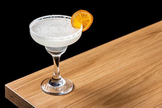 Classic Margarita cocktail in a coupette glass with a salted rim and dried orange slice garnish, on a wooden bar counter