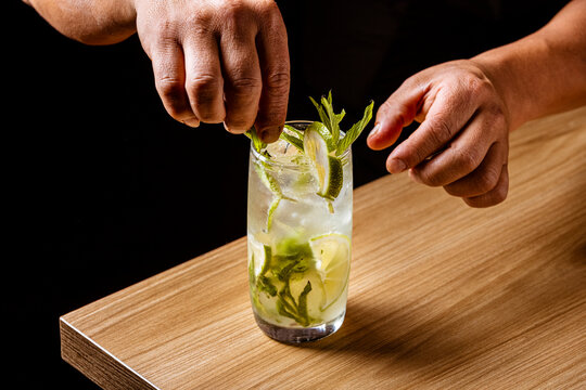 Bartender making a refreshing Mojito cocktail with fresh mint and lime on a wooden bar counter against a black background.