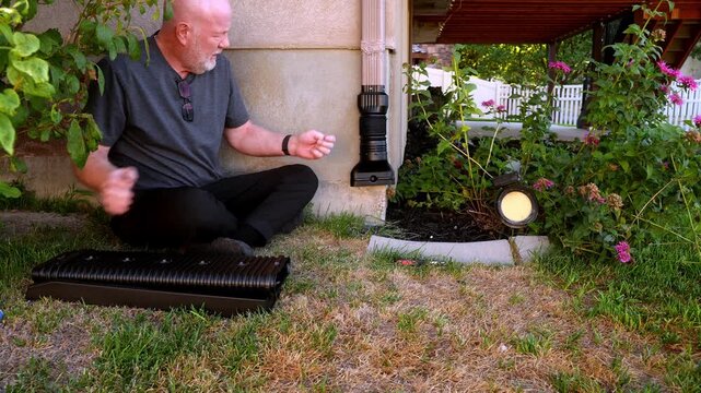 A homeowner sits on the lawn and assembles a black flexible downspout extension beside a residential foundation