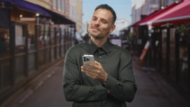 Man holding smartphone, hands visible, smiling while scrolling with crossed arms at a street cafe terrace under red awnings and outdoor seating; contentment.