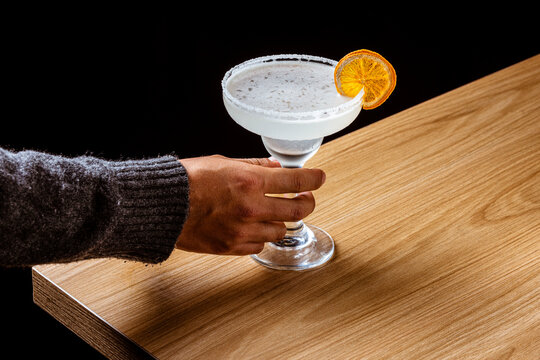 Classic Margarita cocktail in a coupette glass with a salted rim and dried orange slice garnish, on a wooden bar counter