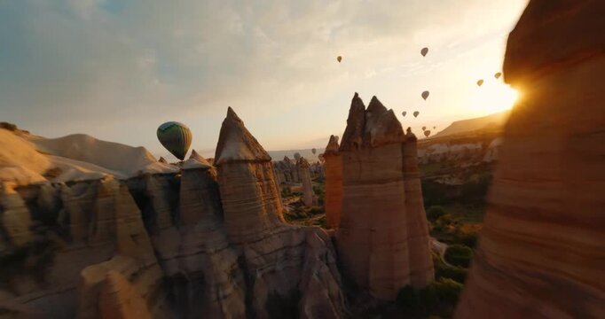 FPV aerial view of numerous hot air balloons drifting over the unique fairy chimneys and rocky landscape of Cappadocia during golden hour in Turkiye.