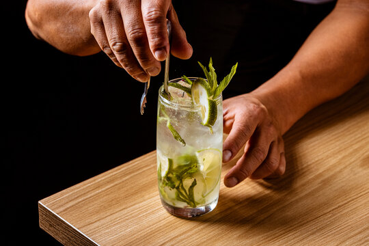 Bartender making a refreshing Mojito cocktail with fresh mint and lime on a wooden bar counter against a black background.