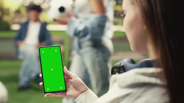 Vertical chroma key appears on an editable smartphone screen held by a young woman outdoors in a university campus study area, offering a green screen phone mockup for mobile screen replacement.