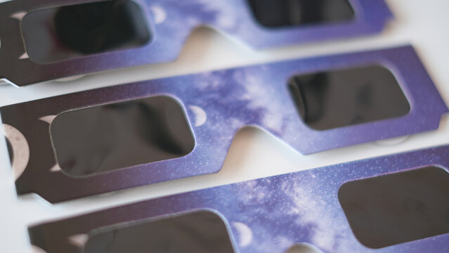 Close-up flat lay view of protective solar eclipse paper glasses featuring a cosmic purple night sky and moon phases design, arranged neatly on a white surface for astronomy and space event concepts.