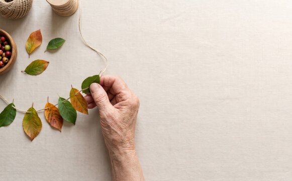 Elderly hands making an autumn leaf garland on rustic fabric background