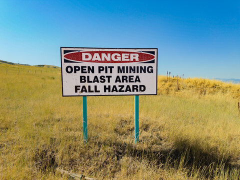View of a danger warning sign for an open pit mining blast area and fall hazard situated in a dry grassy field in North Salt Lake, Utah, United States.