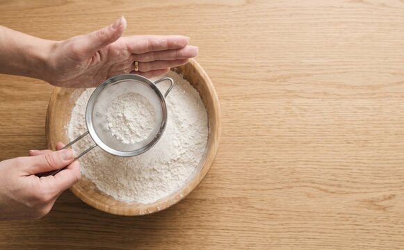 Woman sifting white flour through a metal sieve into a wooden bowl on a table top