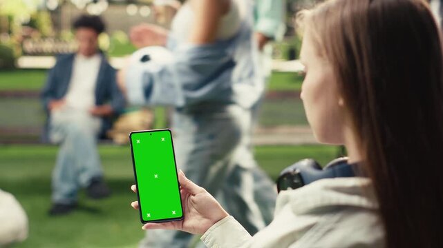 Vertical green screen phone mockup is held close to the camera in an outdoor campus setting, with a blurred student behind and clear space for mobile screen replacement and education content.