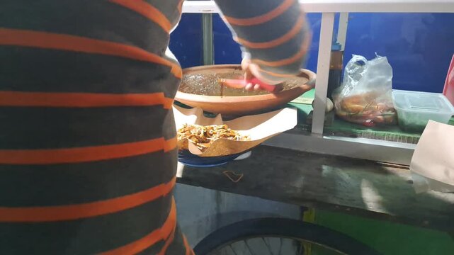 Street food vendor preparing Tahu Tek, a traditional East Javanese dish. Close up of cutting fried tofu, omelet, rice cakes using scissors, mixing rich peanut sauce. SUMENEP, INDONESIA - MAY 27, 2026