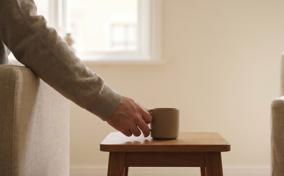Adult man sitting on a sofa and holding a coffee mug on a small wooden table in a living room