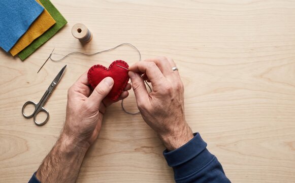 Adult man sewing a red felt heart shape with needle and thread at a wooden table