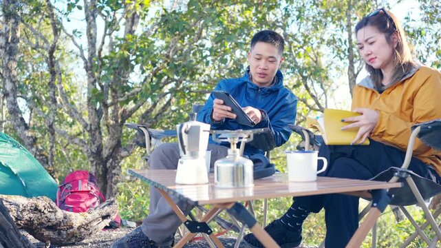 Asian man and woman remote working corporate business on digital tablet and smartphone while travel nature camping in forest with drinking coffee. Digital nomad workation outdoor active lifestyle