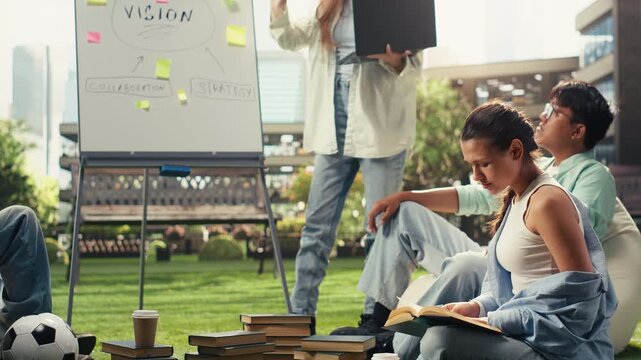 Young woman reads an open book on the grass during a group tutoring session, with a whiteboard and classmates in the background as she studies from a textbook after class on campus.
