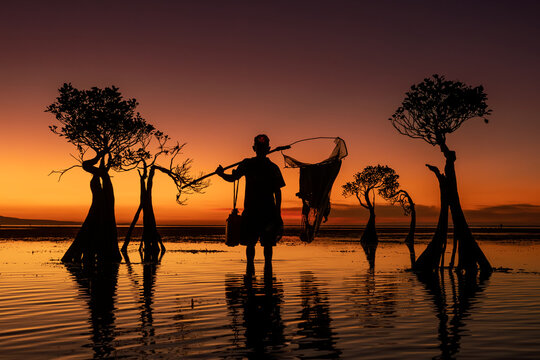 View of a fisherman with a net standing in the shallow water among unique mangrove trees at Walakiri Beach during a vibrant sunset in Kecamatan Pandawai, Nusa Tenggara Timur, Indonesia.