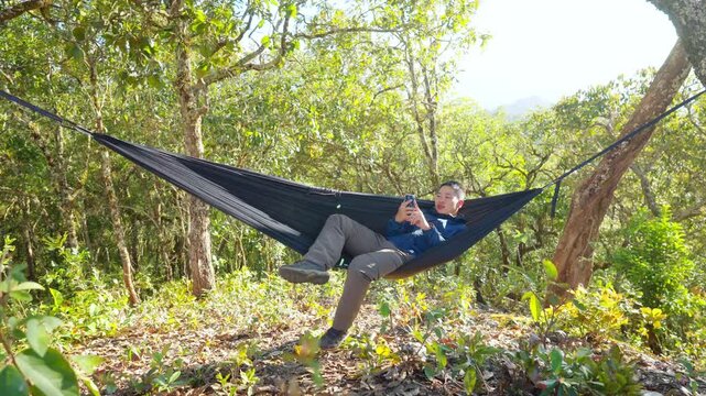 Asian man relaxing in hammock using smartphone while camping in forest enjoy outdoor active lifestyle leisure travel digital connection social media communication, nature healing on holiday vacation.