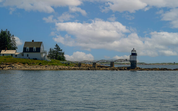 A picturesque lighthouse stands on a rocky shore adjacent to a cozy seaside cottage. The scene is set against a backdrop of blue sky and fluffy clouds.