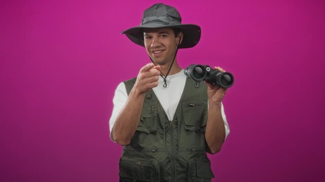 Man in explorer hat and vest points finger to binoculars while holding them in a studio; curiosity exploration adventure.