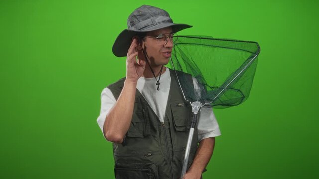 Man fisherman wearing wide brim hat, glasses and fishing vest holds a landing net and cups his ear, smiling against a green screen in studio; curious exploration.