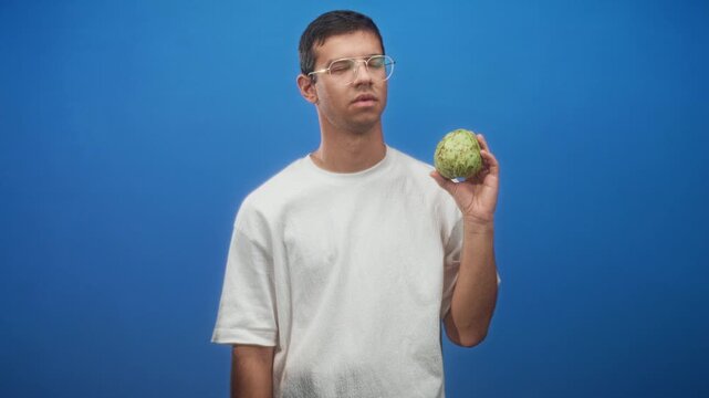 Man holds custard apple in right hand against blue studio backdrop, wearing white tshirt and glasses with a skeptical expression; uncertainty curiosity.