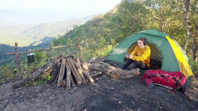 Asian woman listening to music with headphones and using smartphone mobile app while camping on the mountain. People enjoy outdoor active lifestyle nature therapy and wellness relaxing forest bathing