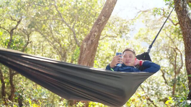 Asian man relaxing in hammock using smartphone while camping in forest enjoy outdoor active lifestyle leisure travel digital connection social media communication, nature healing on holiday vacation.