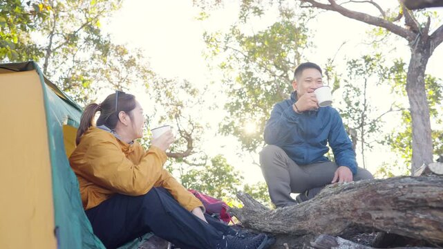 Happy Asian couple drinking hot coffee while travel nature and camping at forest mountain. Man and woman relaxing at campsite, enjoying morning beverage with outdoors active lifestyle nature therapy.