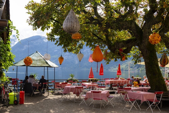 View of an outdoor cafe terrace with red checkered tables and hanging lanterns under a large tree overlooking the lake and mountains in Hallstatt, Upper Austria, Austria.