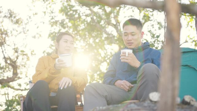 Happy Asian couple drinking hot coffee while travel nature and camping at forest mountain. Man and woman relaxing at campsite, enjoying morning beverage with outdoors active lifestyle nature therapy.