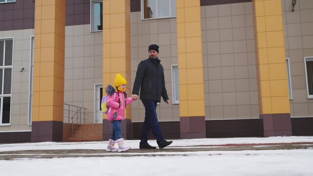 girl holding her father hand, school road education classes, concept happy family, little child kid daughter with school backpack walking through snow boots, school student with backpack, daughter bag