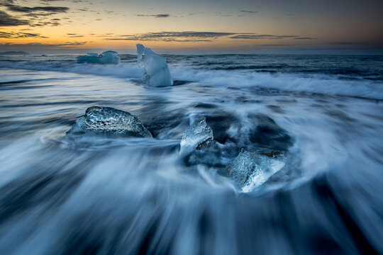 View of ice chunks from Jokulsarlon Glacier Lagoon washing up on the black sand beach at sunset with long exposure waves swirling around the ice Hornafjordur, Iceland.