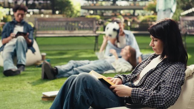 Woman student sits on a beanbag holding an open book and studies alone outdoors with a notebook, supporting independent learning in a modern campus setting with a softly blurred background.
