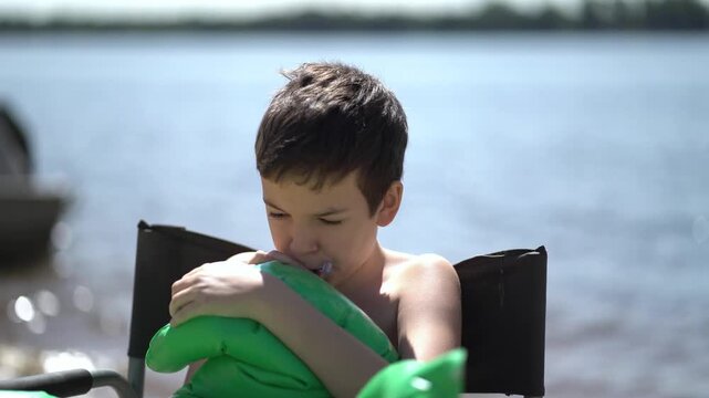 Young boy sits in a chair by the water and carefully inflates a green swimming mattress with his mouth on a sunny summer day at the beach.