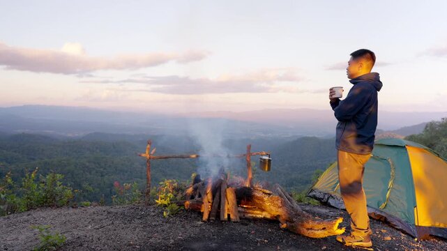 Asian man drinking hot coffee while solo travel nature and camping at forest mountain. Male backpacker relaxing at campsite, social detox, enjoy nature therapy morning beverage with outdoor lifestyle