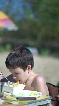 Young boy hungrily slurps ramen noodles outdoors on the beach during a summer picnic, capturing an authentic childhood moment in bright natural light.