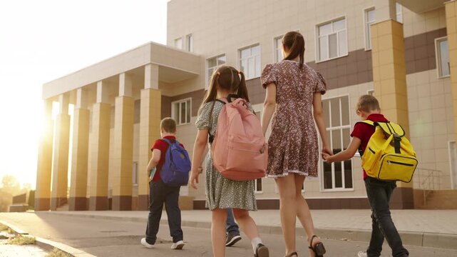 School entrance with students and parents, Sunny day at modern school building, Pupils going towards classroom with guardians, Back-to-school morning scene, Rear view of children with backpacks
