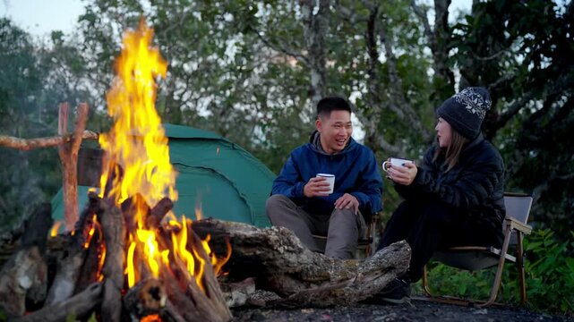 Happy Asian couple drinking hot coffee while travel nature and camping at forest mountain. Man and woman relaxing at campsite, enjoying morning beverage with outdoors active lifestyle nature therapy.