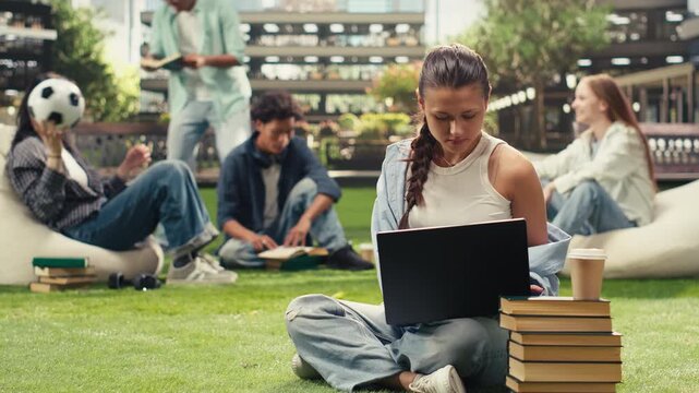 Smiling young woman works on a laptop from the grass at a modern campus and types coursework notes, with a backpack and stacked books nearby in an independent outdoor study session.