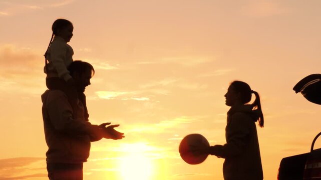 Family playing with ball in nature at sunset, silhouettes. Daughter sits on father's shoulders, throwing ball with mother. Family travel adventures by car, active leisure. Weekend road trip to nature.