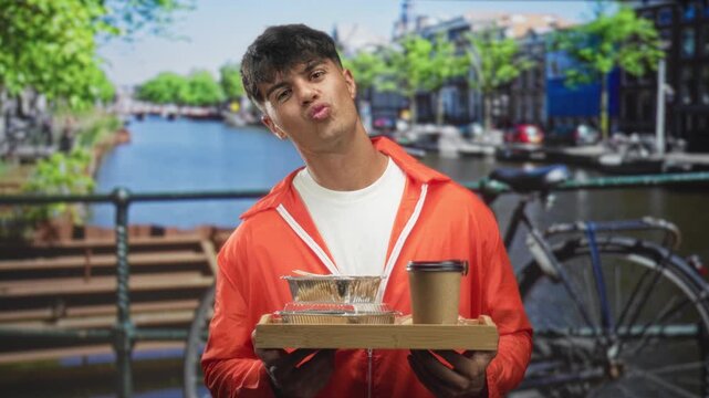 Young man in orange prisoner jumpsuit holds a breakfast tray, lips puckered in a kiss gesture, hands gripping tray in a studio; mischievous defiance.