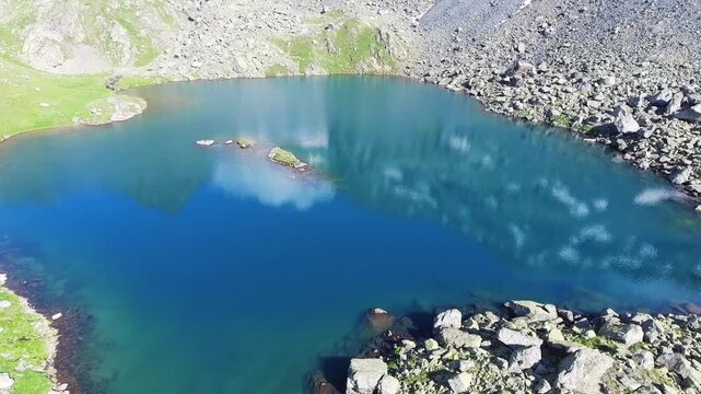 Aerial drone view of alpine tarn lake with rocky shoreline and green meadow in mountain terrain.