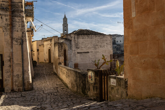 View of a narrow cobblestone street lined with historic stone buildings and a bell tower in the background under a clear blue sky in Matera, Basilicata, Italy.