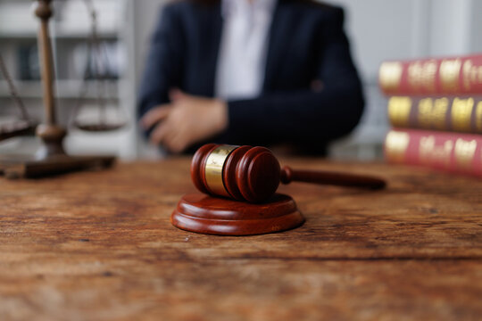 Gavel resting on a wooden desk with a blurred legal professional, scales of justice, and law books in the background
